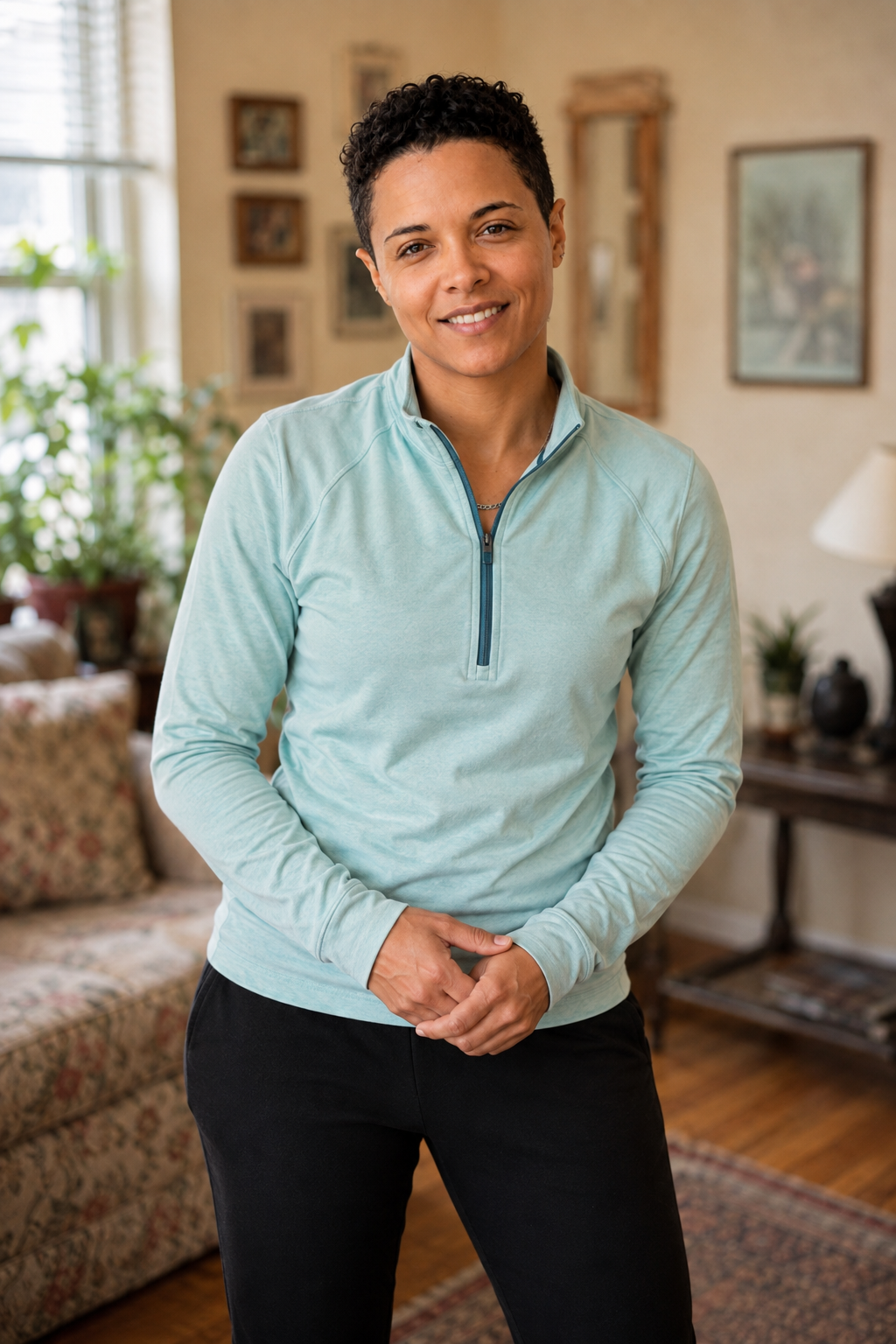 A person in athletic clothing stands in a home living room, hands loosely clasped, looking toward the camera.